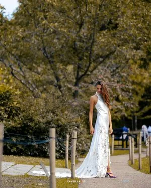A white lace maxi dress is worn outdoors on a path with autumn trees and wooden posts in the background.