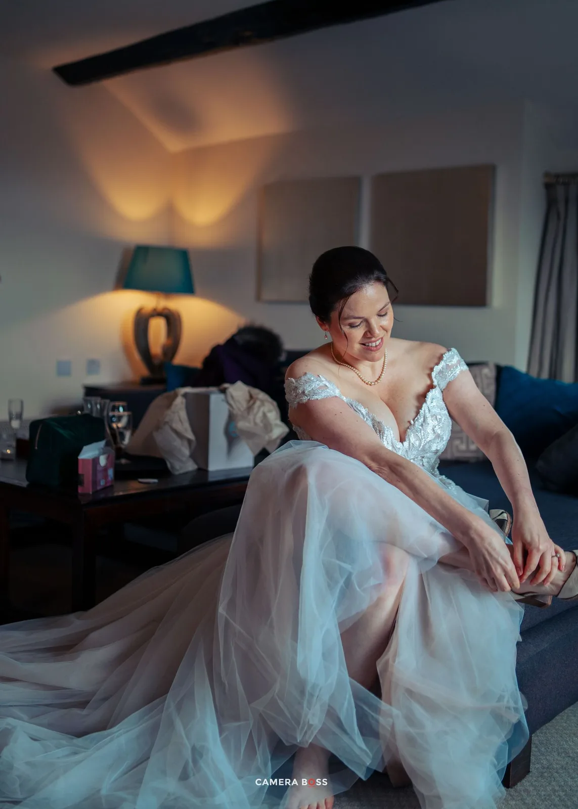 A bride in a flowing white lace gown sits on a couch while adjusting her shoe in warm ambient lighting.