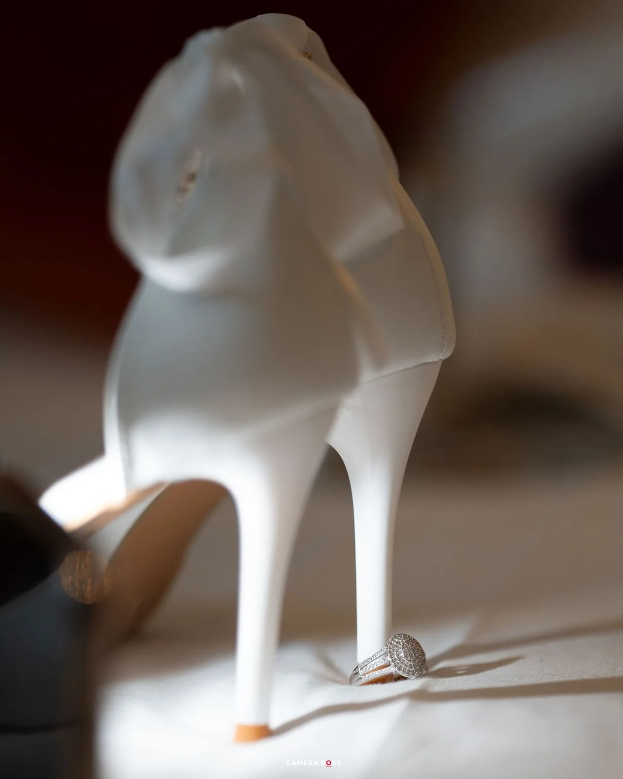 An elegant white satin high heel wedding shoe with ring displayed, photographed against a blurred dark background.