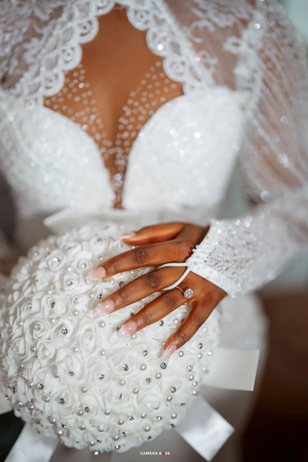 Close-up detail of a beaded white wedding dress bodice with lace details and a bride's hands showing her ring.
