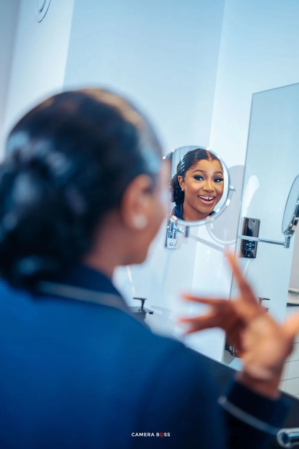 A person smiles while looking in a bathroom mirror, wearing professional attire in a bright blue setting.