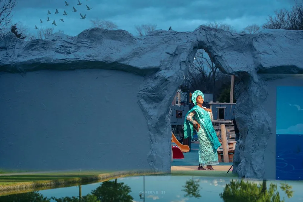 A figure in a teal dress stands in a stone archway against a dramatic stormy sky with mountains in the background.