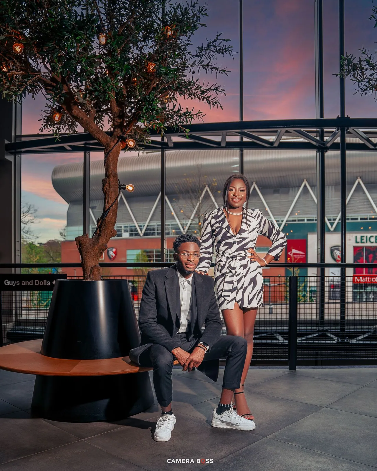 Stylish couple poses at sunset outside modern building with geometric architecture and decorative tree.