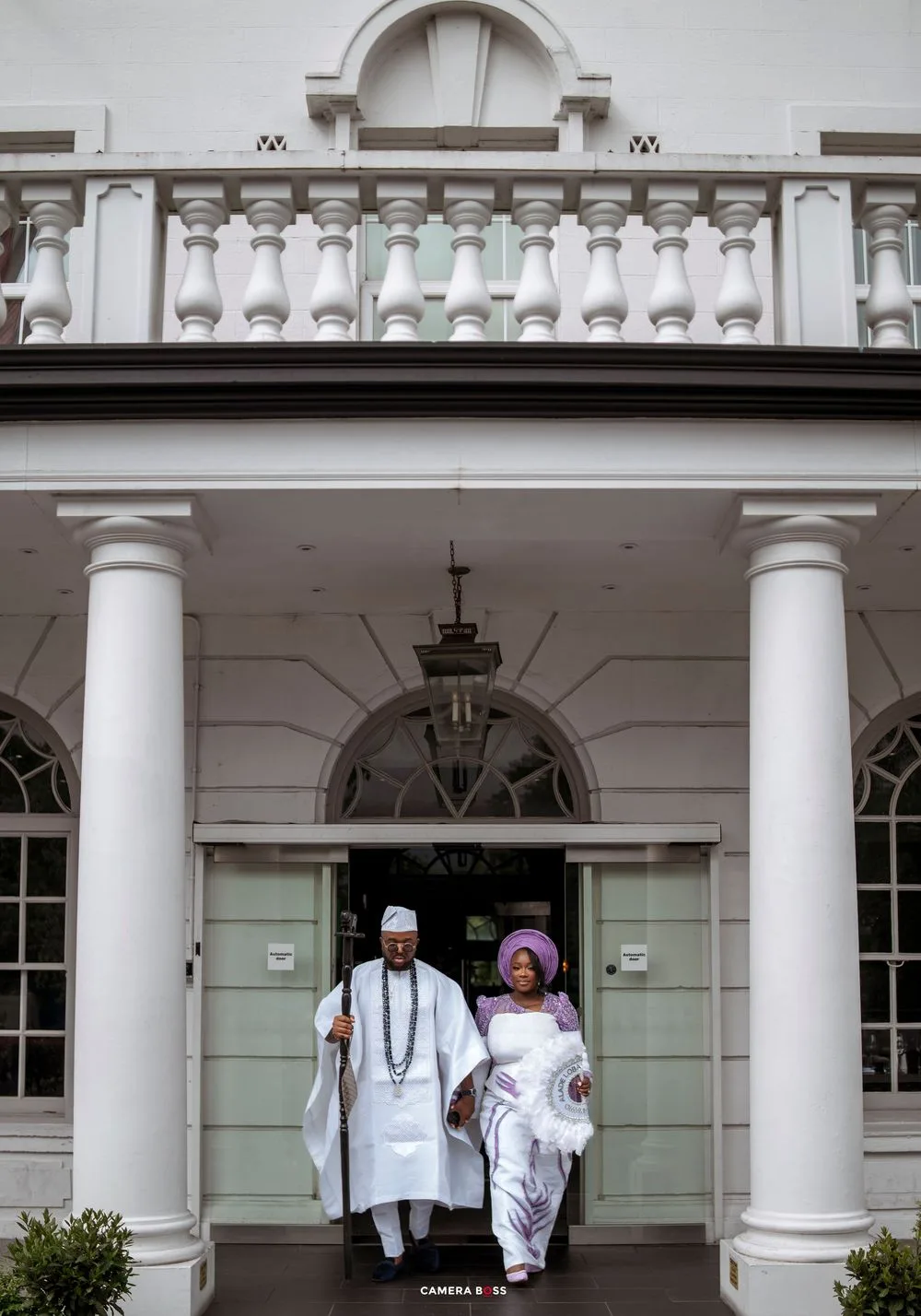 Nigerian couple in traditional Yoruba attire exit elegant white colonial building with columns during wedding ceremony.