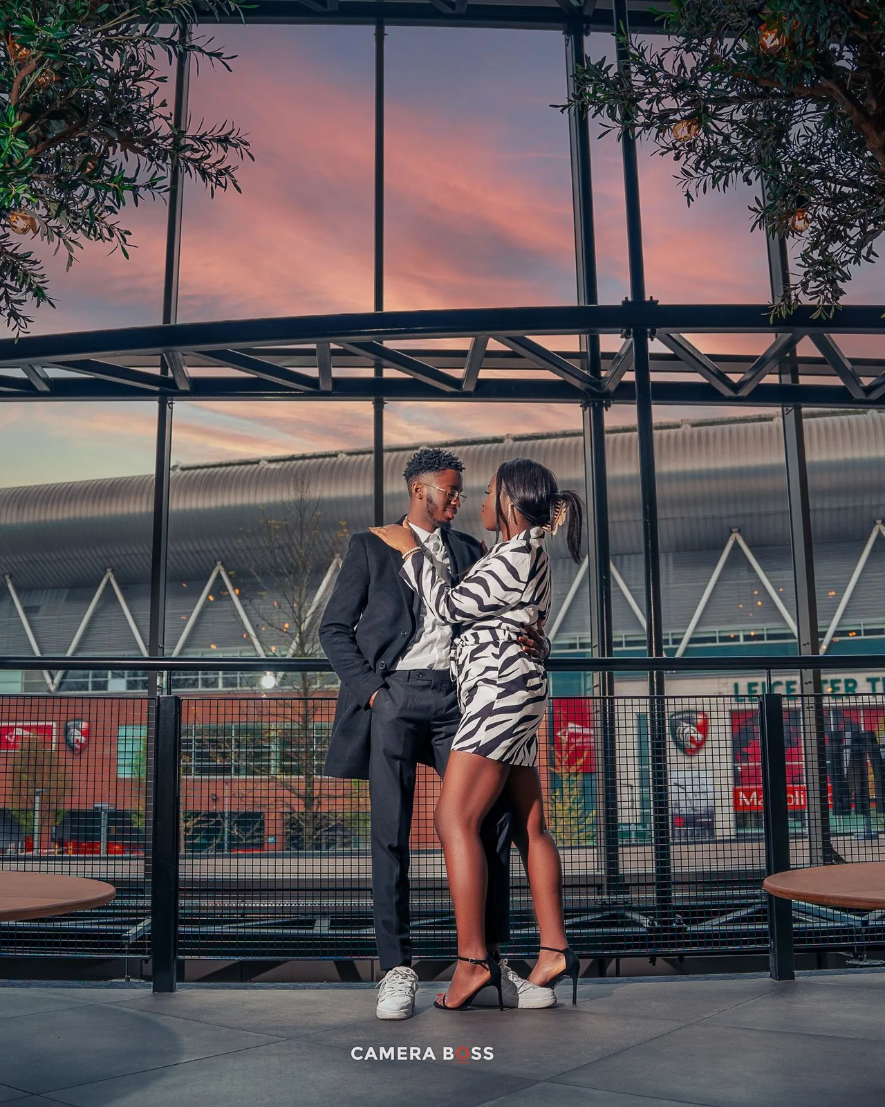 A couple shares an intimate moment outside a modern stadium building at sunset with pink and orange skies.
