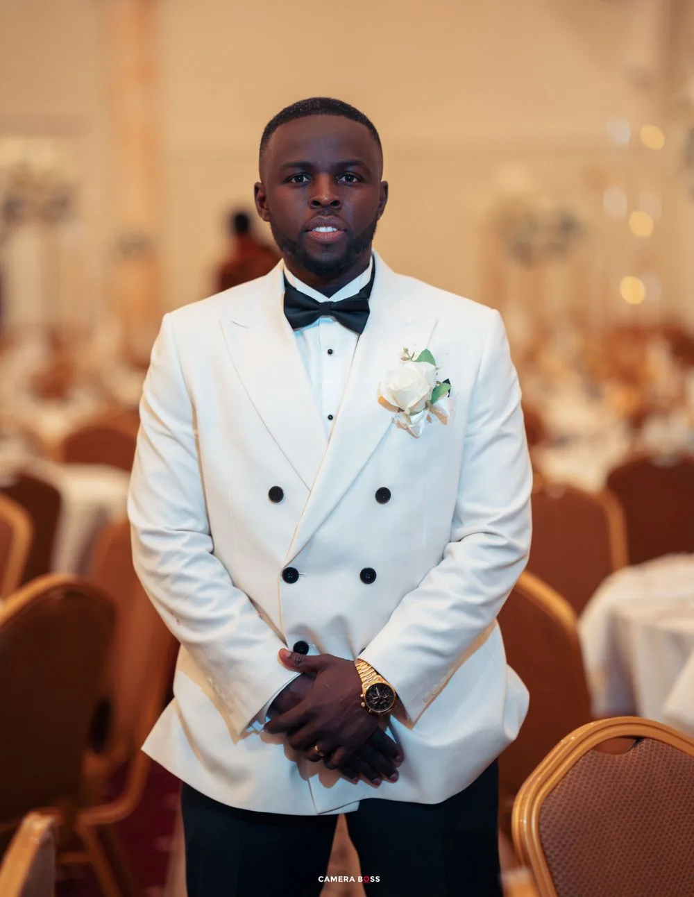 A person in a crisp white double-breasted tuxedo jacket with black bow tie and boutonniere stands in an elegant ballroom.
