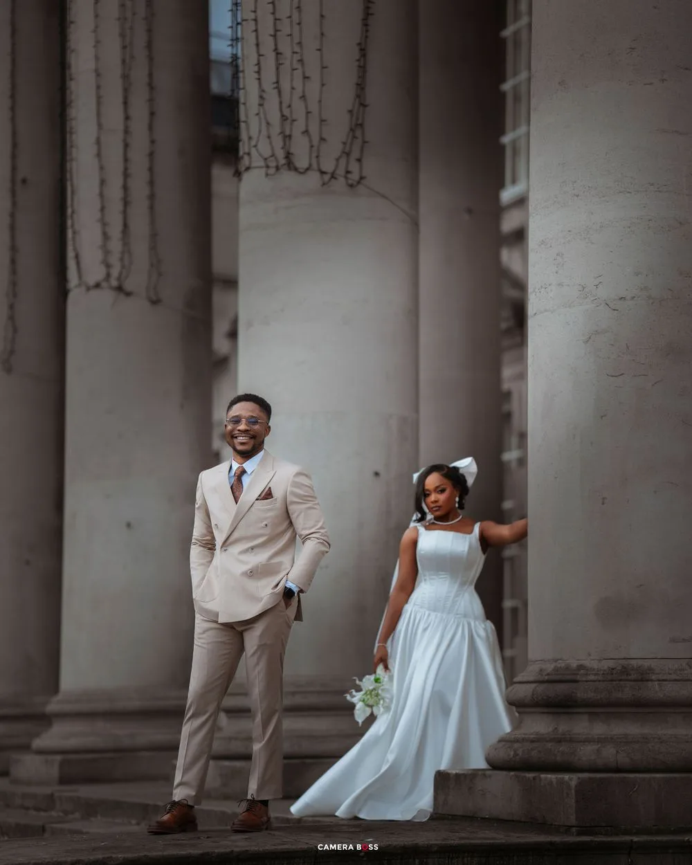 A couple in formal attire shares playful moments between elegant stone columns during a romantic photoshoot.