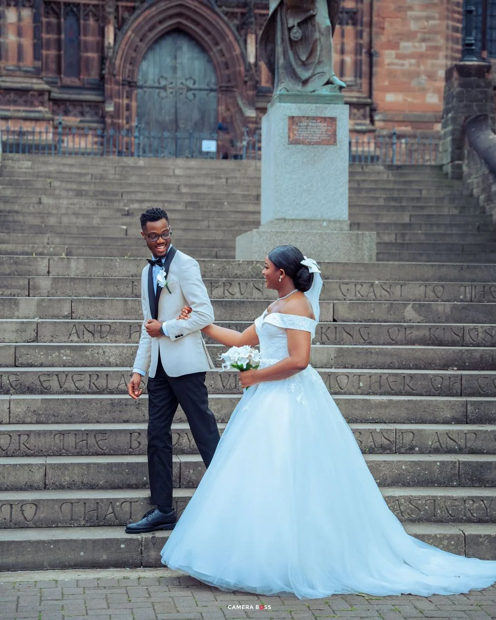 A couple in formal wedding attire share a moment on stone steps in front of a historic church entrance.
