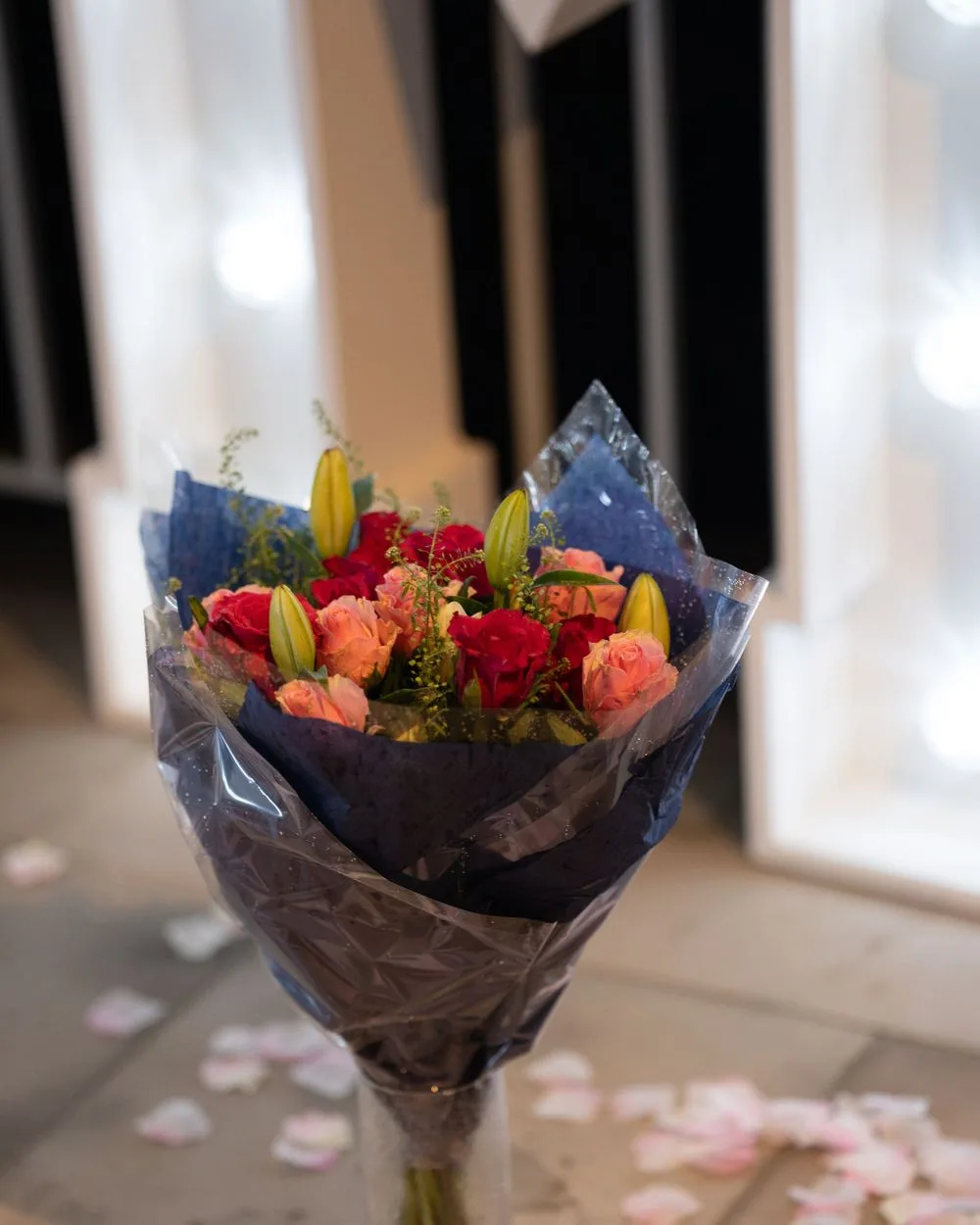 A vibrant bouquet wrapped in navy blue paper features red and pink roses with yellow lilies against a soft background.