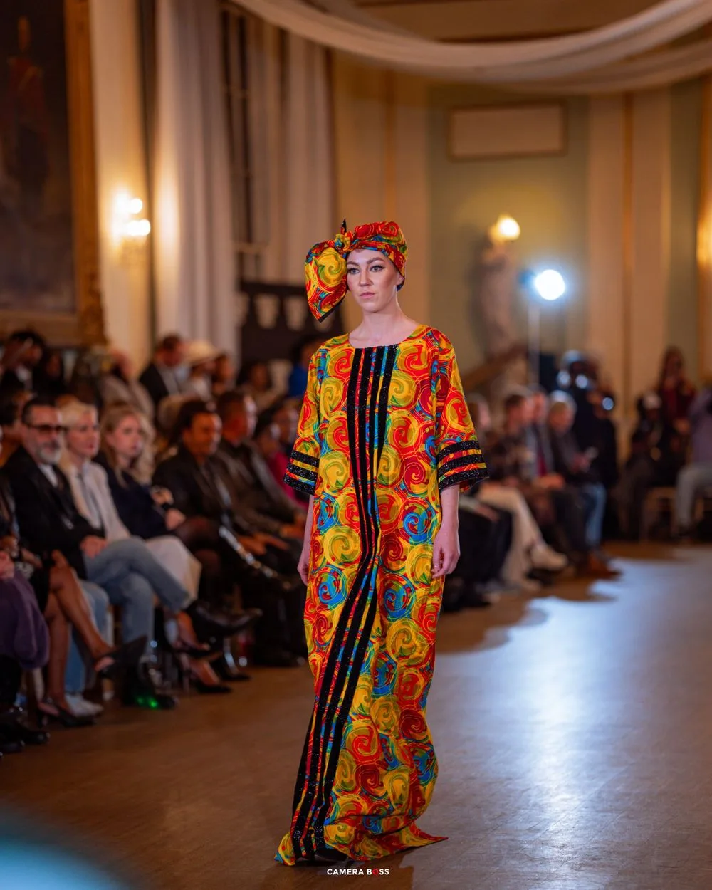 A high-fashion runway show featuring a bright floral print dress with orange and yellow blooms against dark stripes.