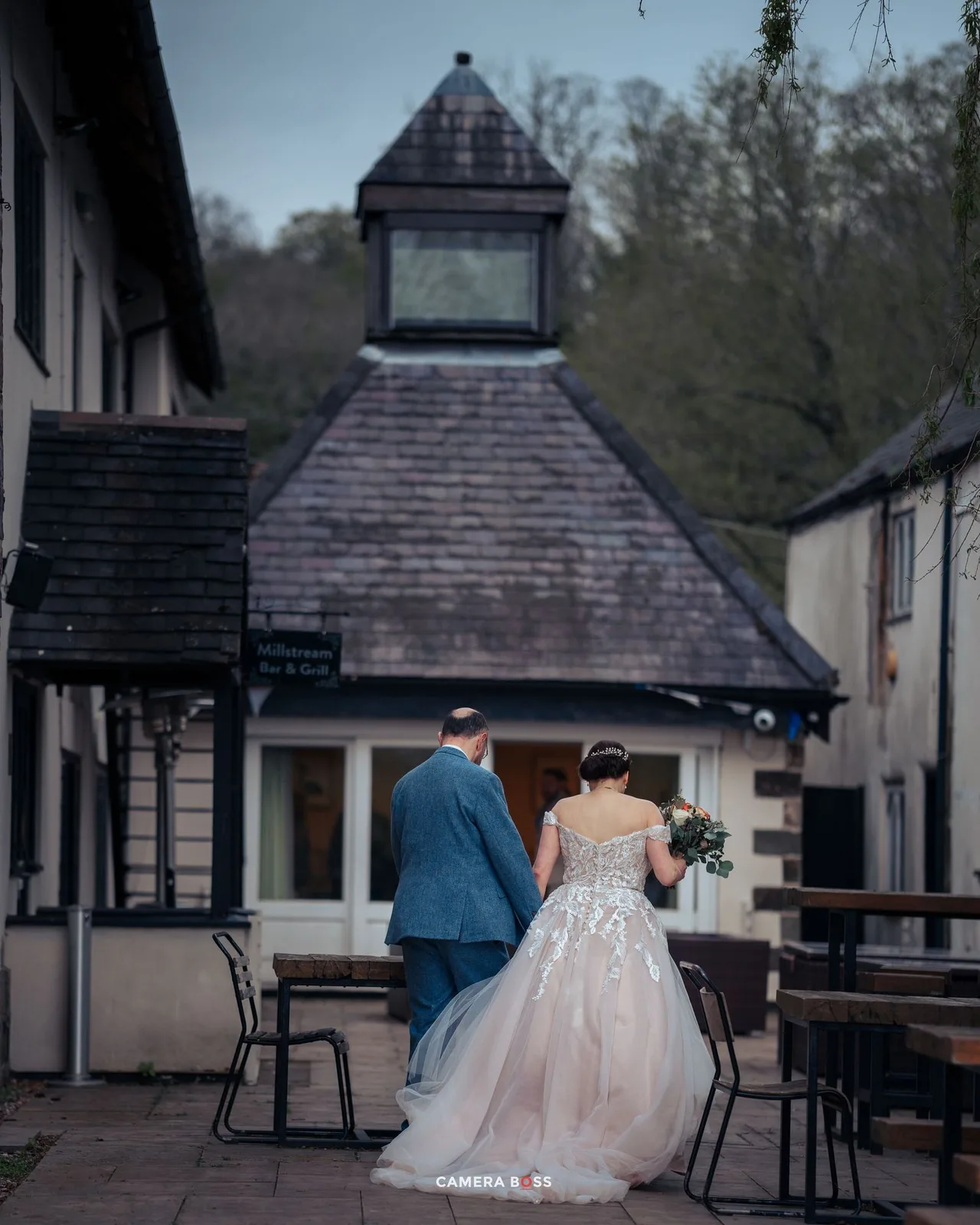 A couple in wedding attire stand together on a patio with outdoor seating and a rustic gazebo-style structure in the background.