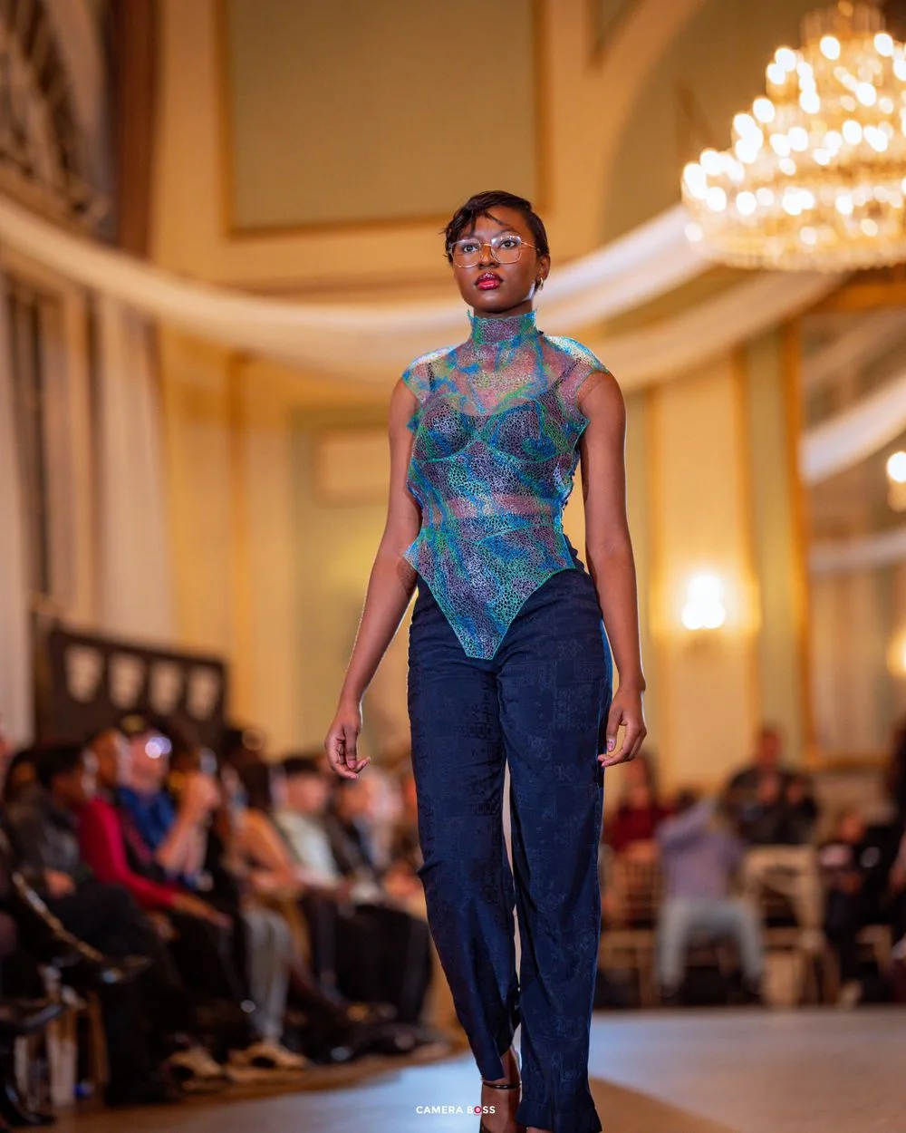 Fashion model walks runway in elegant blue sheer mesh top and denim pants beneath ornate chandelier at fashion show.