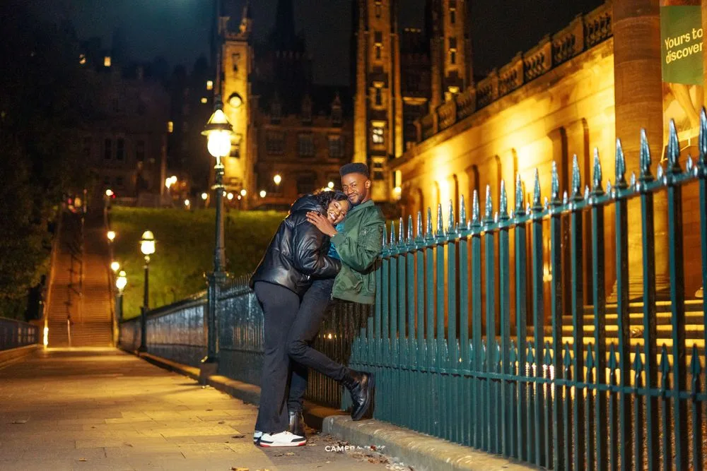 A photographer takes a nighttime photo through a blue iron fence with illuminated buildings in the background.
