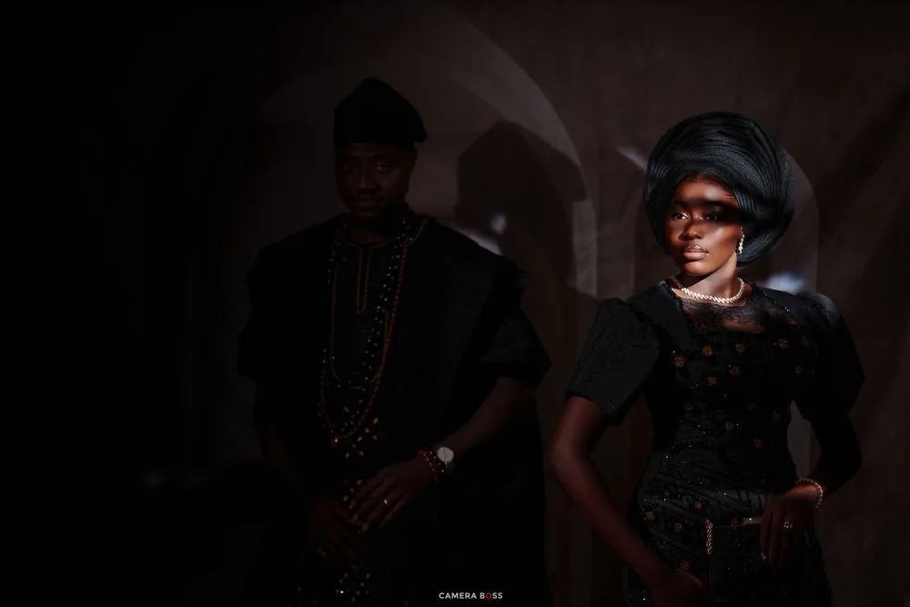 A Nigerian couple in elegant traditional attire pose dramatically in dim lighting, showcasing cultural fashion.