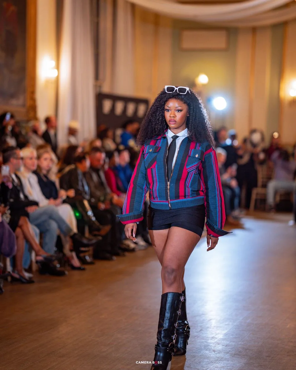 Model struts down catwalk wearing colorful bomber jacket, shorts and boots during indoor fashion show presentation.