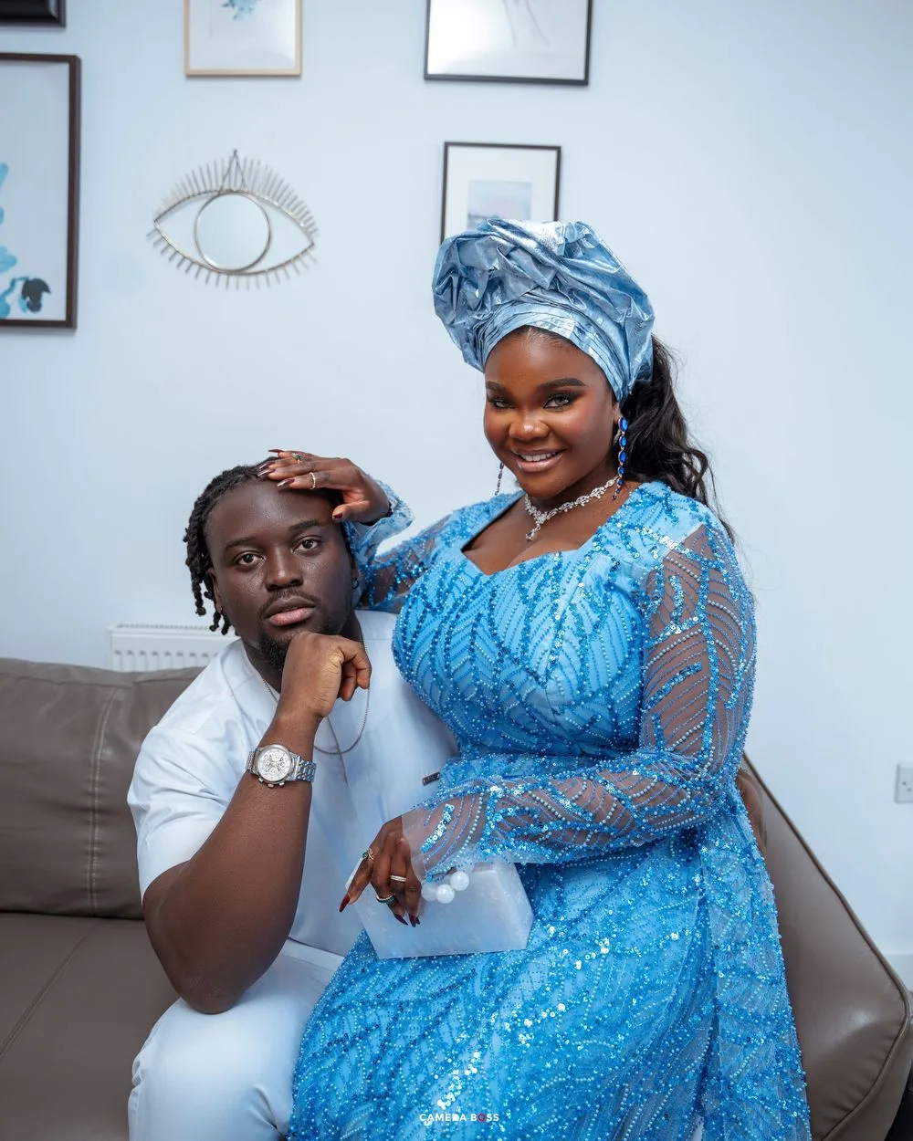 Two people pose together on a couch, one wearing an elegant blue lace dress with matching headwrap, the other in white attire.