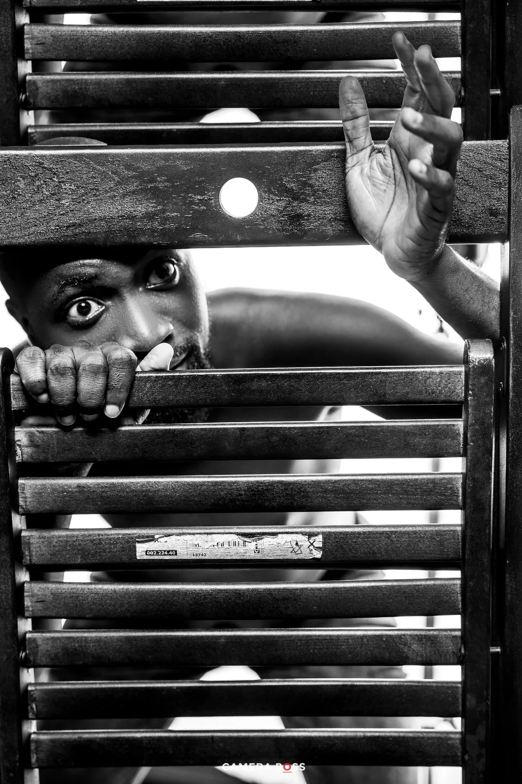 A dramatic black and white photograph showing eyes peering through horizontal metal bars or shutters.