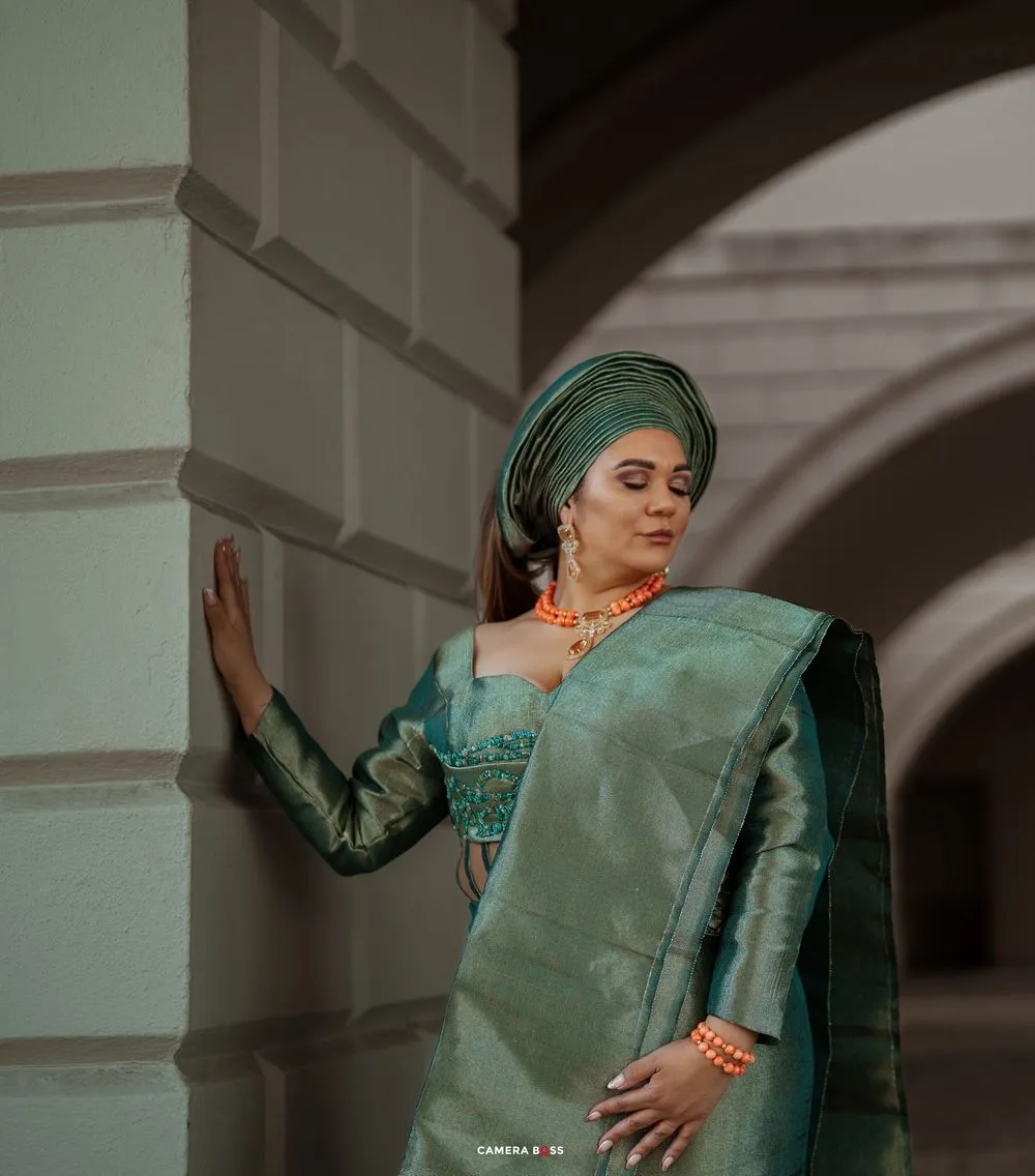 Woman in emerald green headscarf and dress poses gracefully against architectural backdrop in moody lighting.