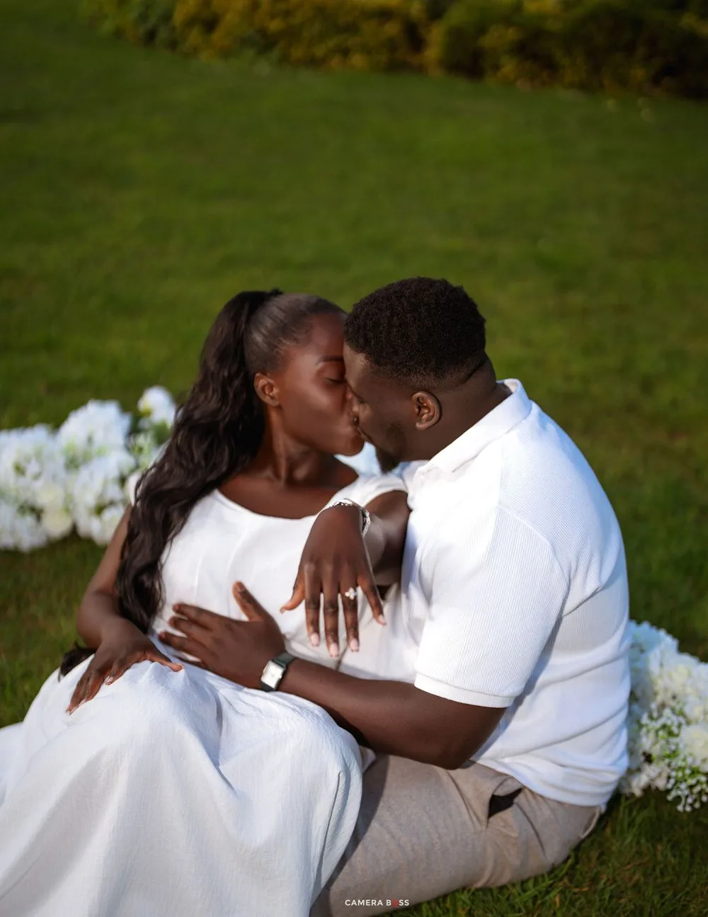 A couple shares a romantic kiss while sitting on green grass beside white flowers during an outdoor engagement photo session.