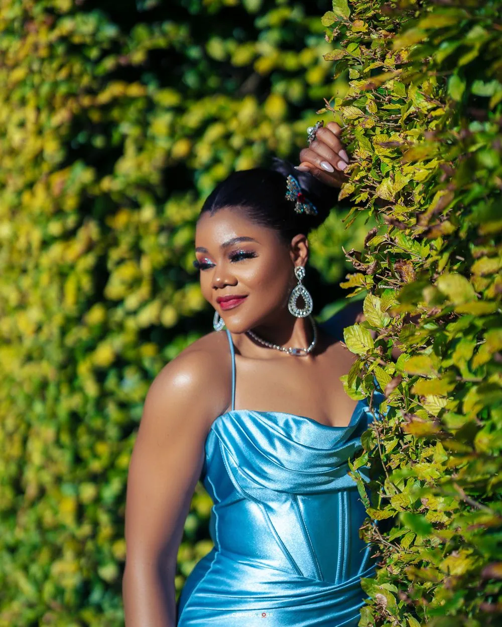 A woman in a light blue satin formal dress poses elegantly against a lush green hedge backdrop.