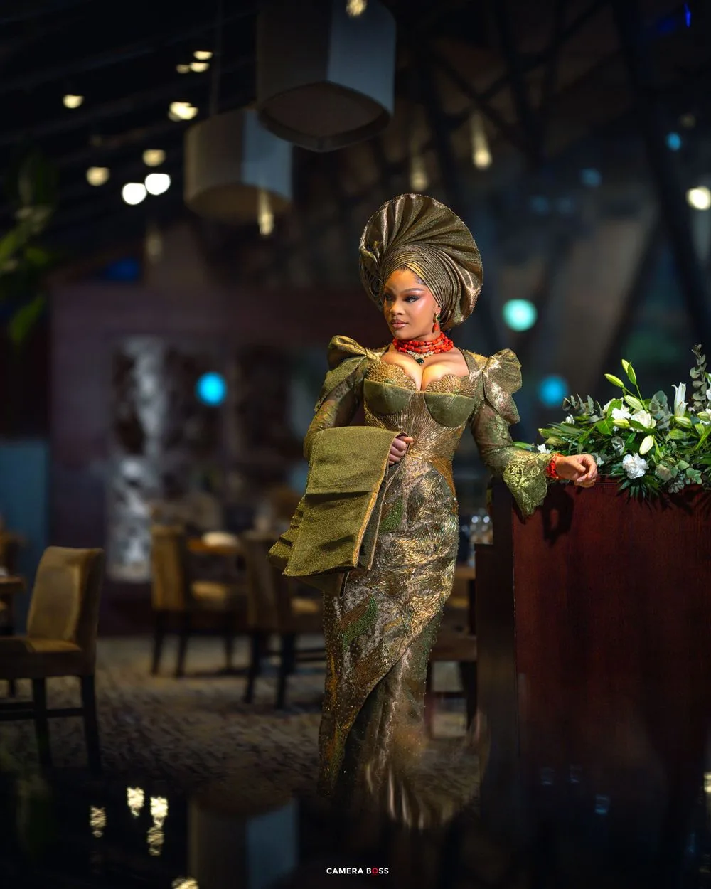 Fashion model in elaborate green kebaya dress with gold headdress poses elegantly at an upscale restaurant setting.