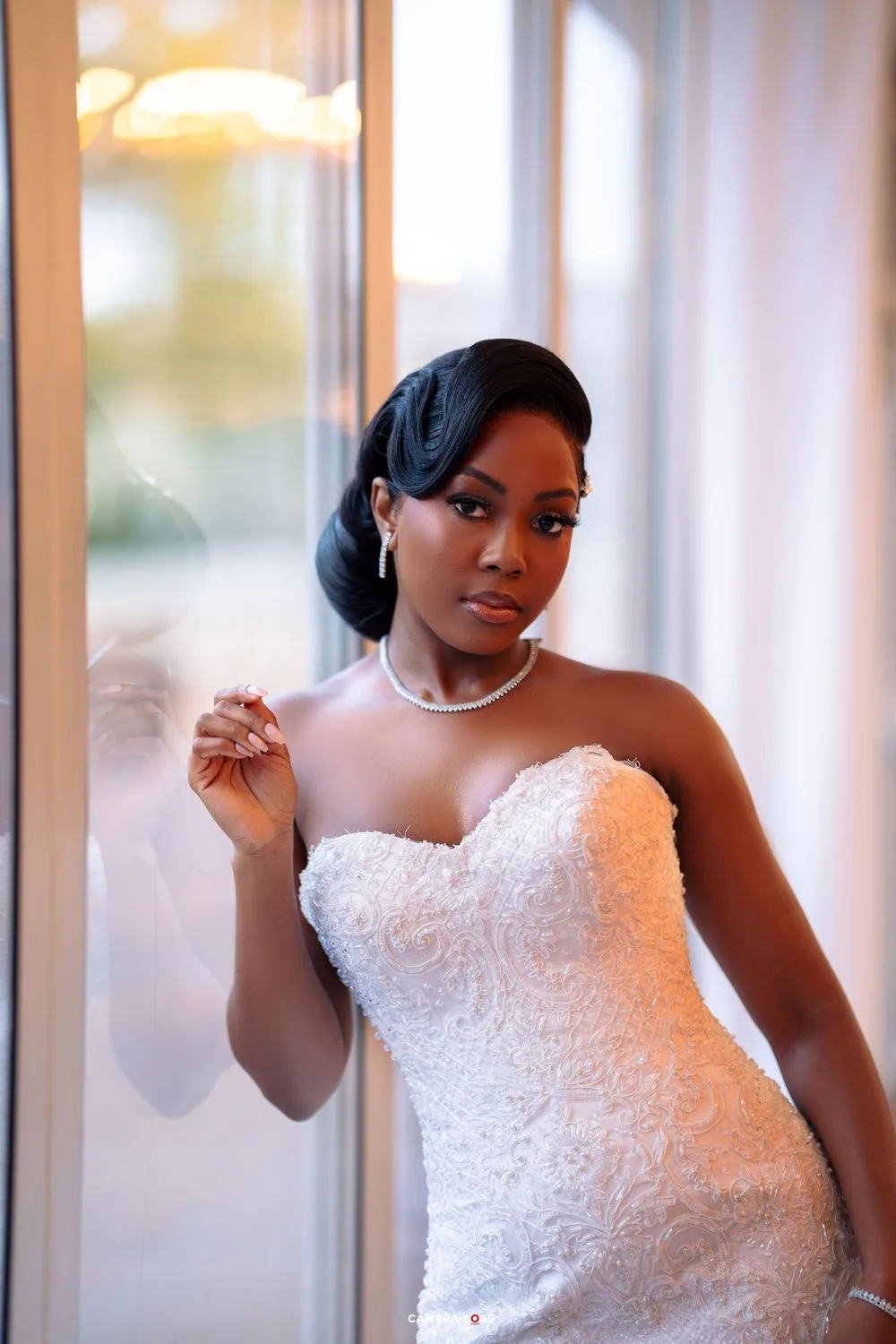 Beautiful bride in a white lace strapless wedding gown and pearl necklace poses elegantly by a window.