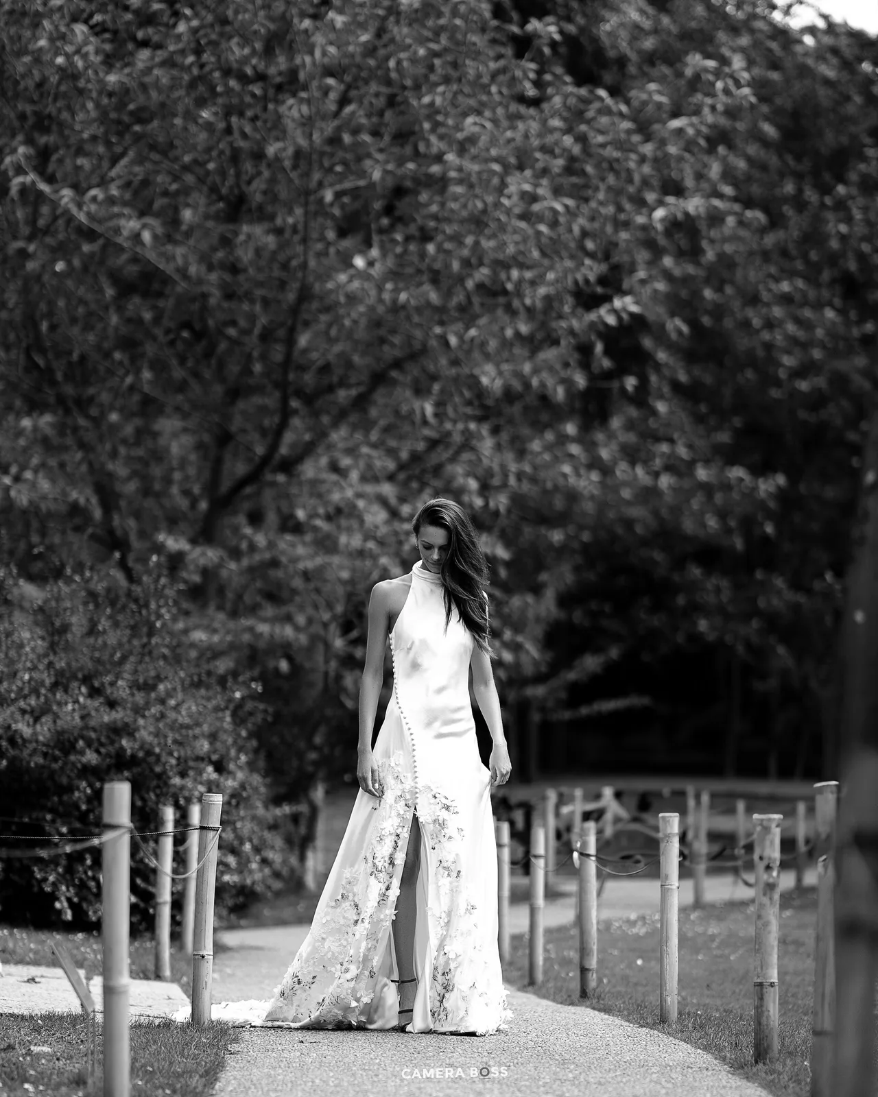 A black and white photograph of a bride in a flowing lace gown standing on a path lined with wooden posts.