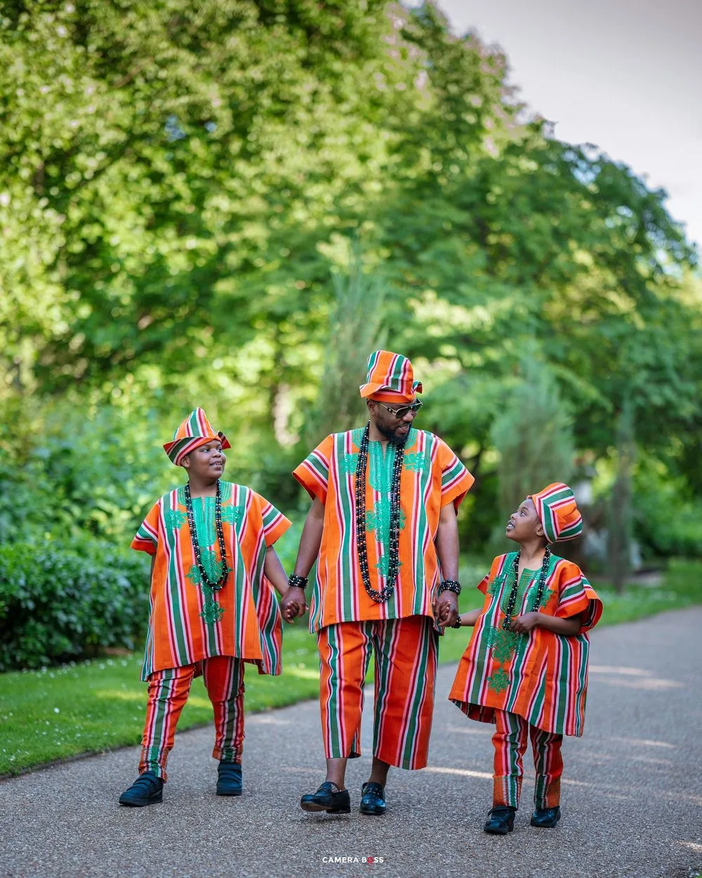 Family holds hands while walking outdoors in matching orange and green African traditional clothing.