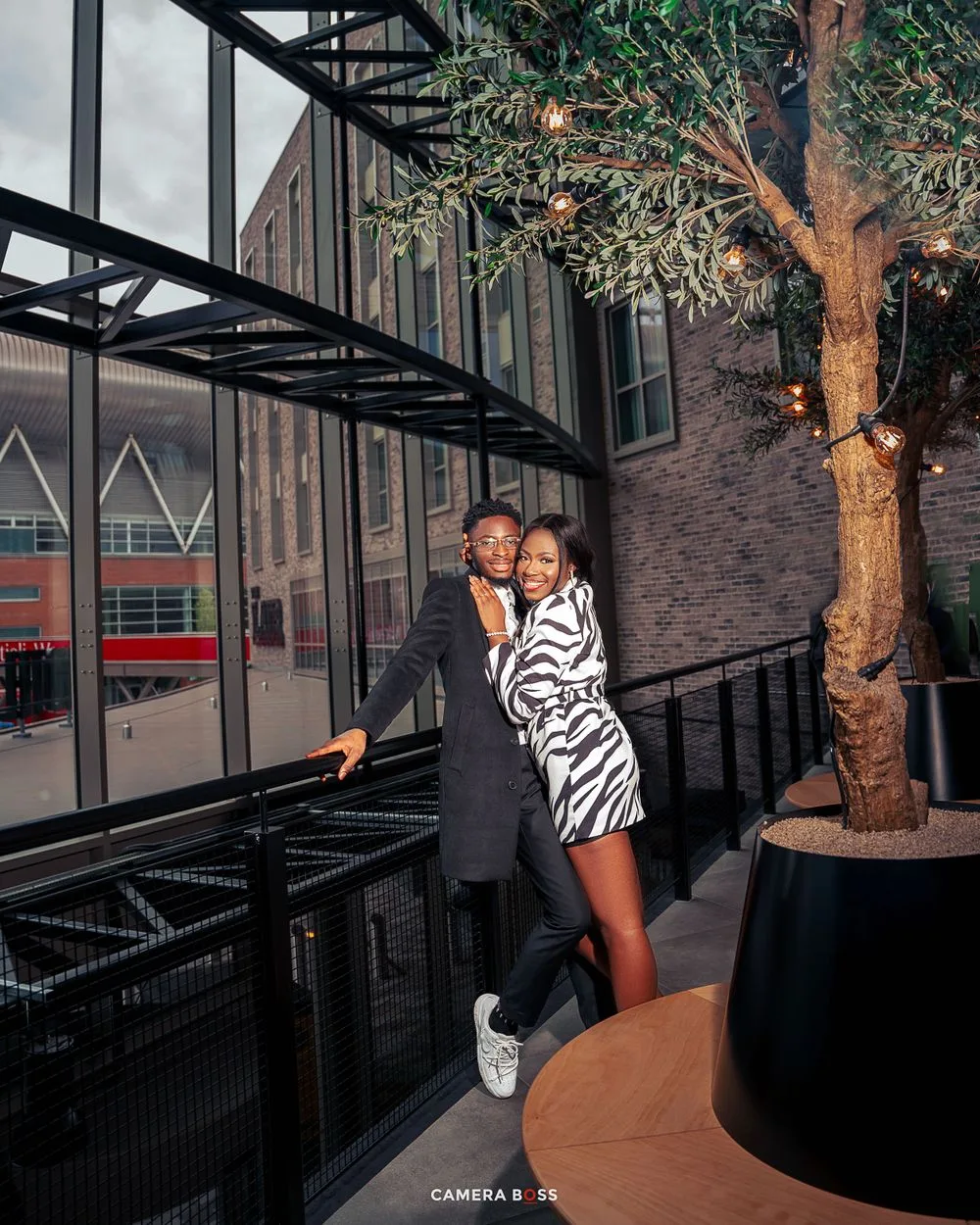 A couple shares a romantic moment outside a modern building with glass windows and a decorative tree at night.