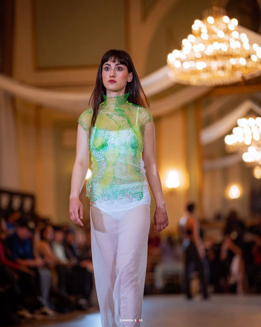 Model wearing sheer mint green qipao-inspired dress with white pants walks runway under elegant chandeliers.