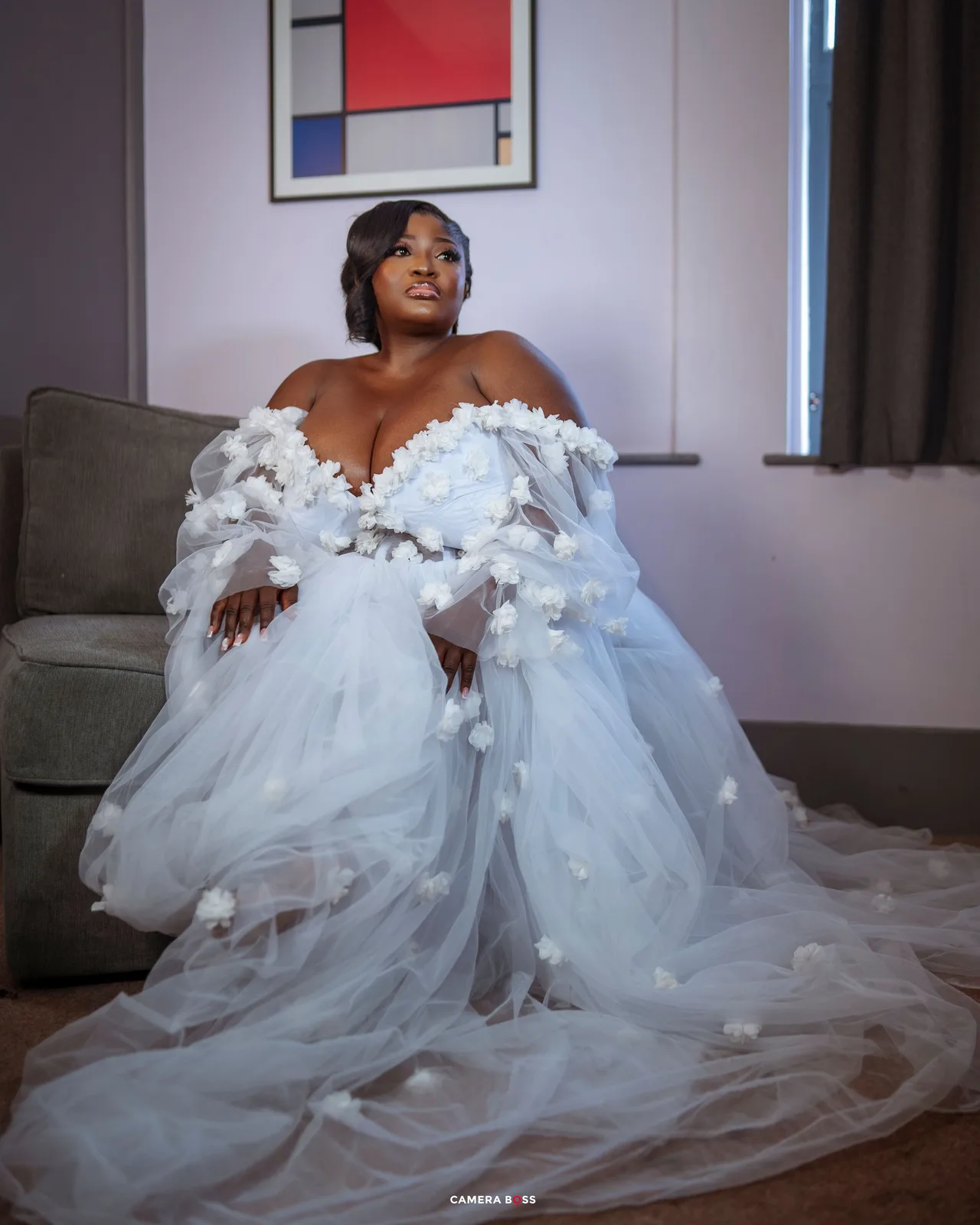 A stunning off-shoulder wedding gown with delicate floral appliques and flowing tulle skirt is showcased in a living room setting.