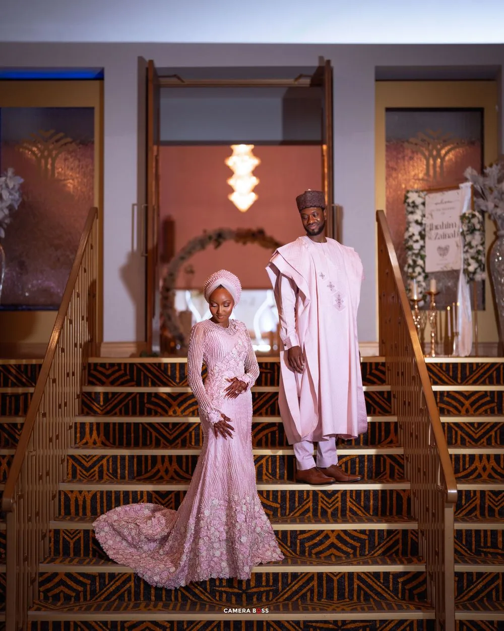 A couple in matching soft pink traditional formal attire pose elegantly on grand wooden stairs at an event venue.