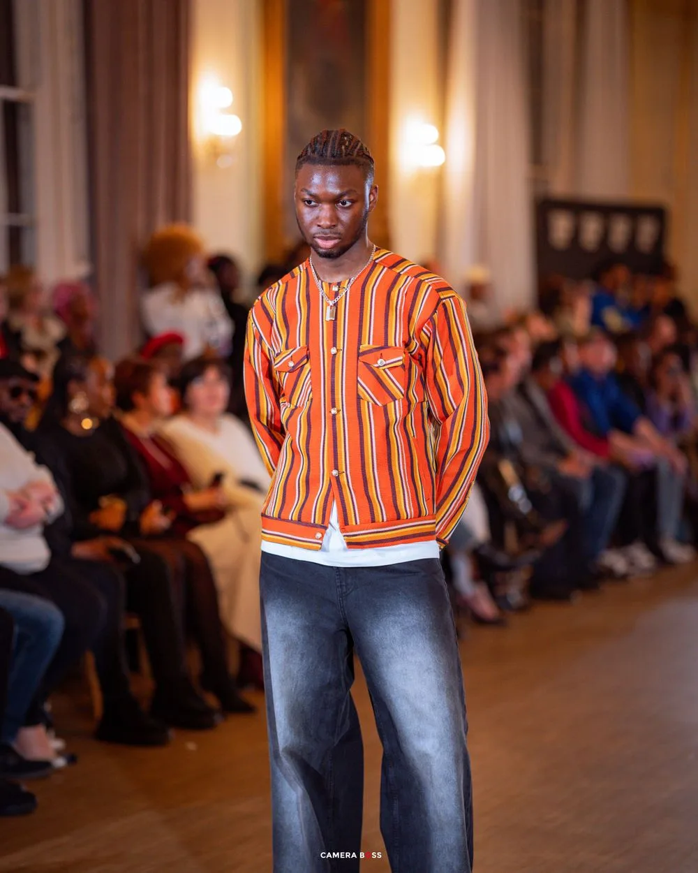 Model walks runway wearing orange striped jacket with denim jeans at fashion show with audience in background.