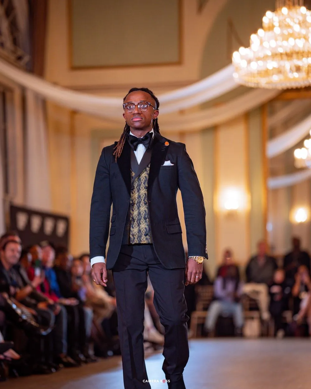 A model walks the runway in a formal navy suit with patterned vest at an elegant event with chandeliers and draped decor.