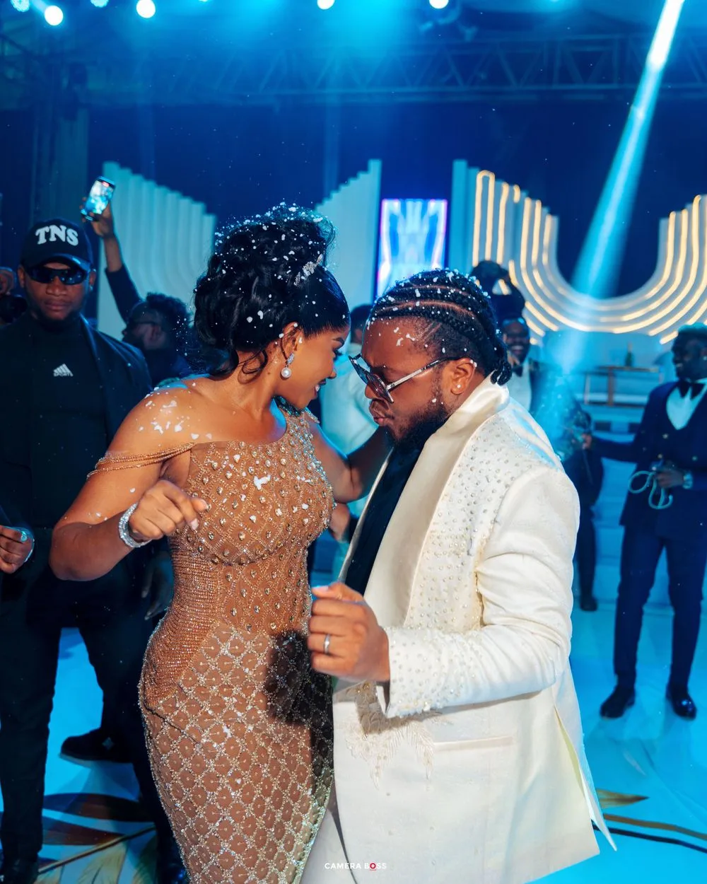 Two people dancing at an elegant blue-lit event, with one wearing a sparkly nude gown and the other in a white suit.