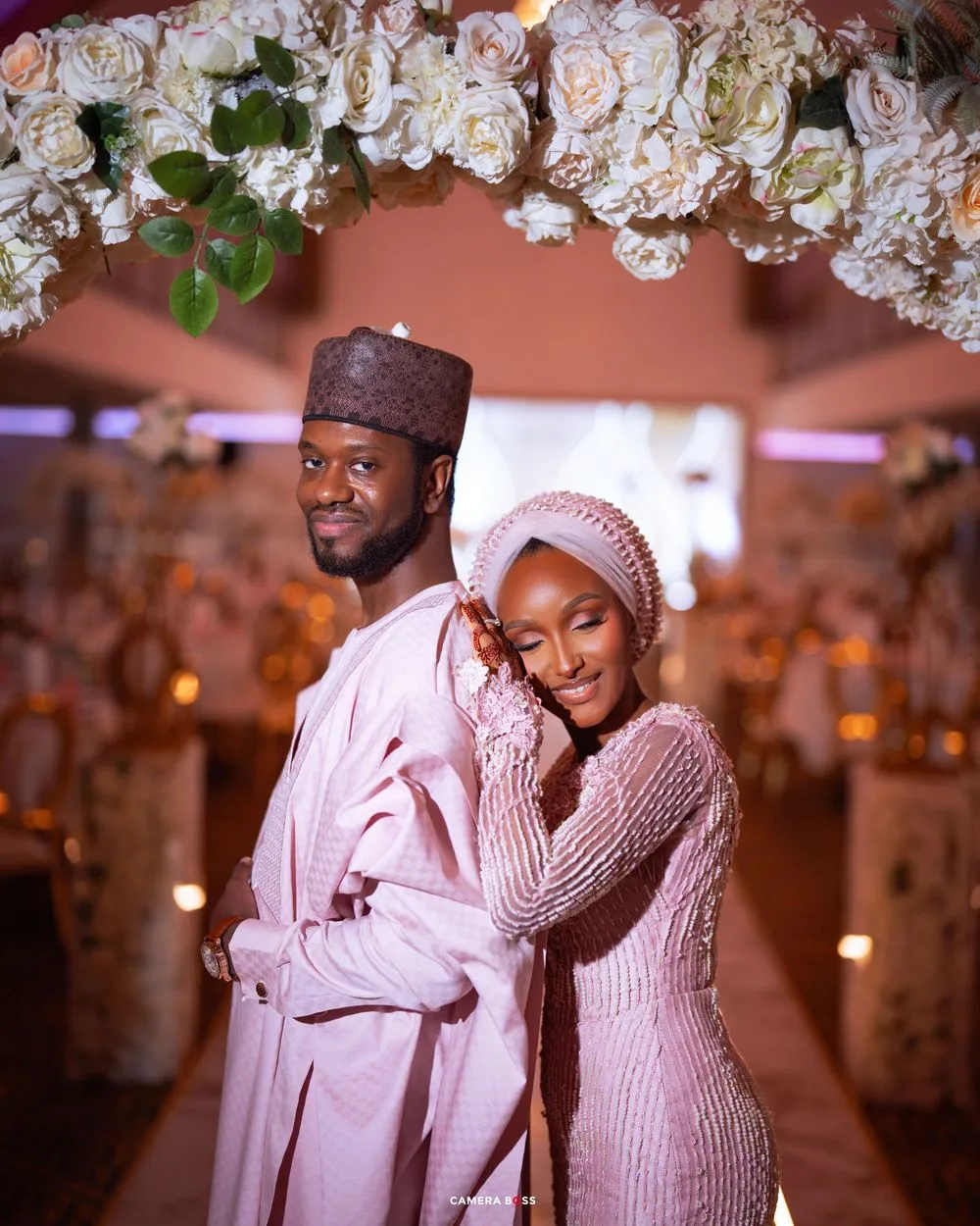 A couple in matching white traditional attire embraces under an elegant wedding arch adorned with white flowers.