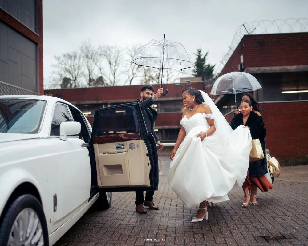 A Rolls Royce awaits as people gather around a car door on a rainy day, with one wearing an elegant white dress.