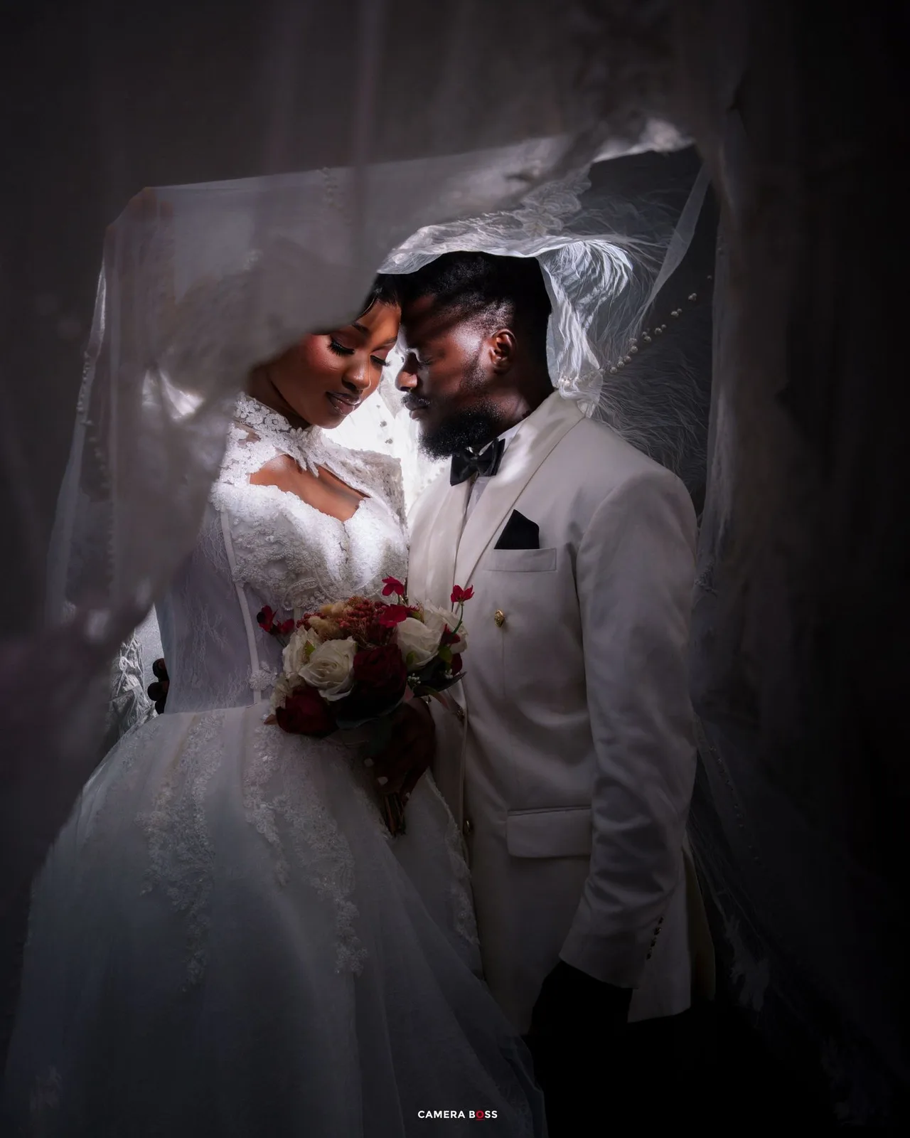 Bride and groom share romantic moment under wedding veil, groom in white suit, bride holding red and white bouquet.