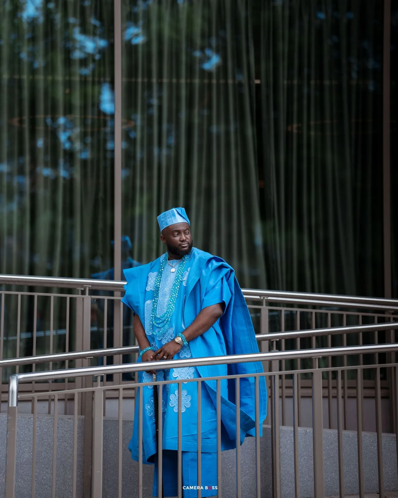 Person wearing vibrant blue traditional African agbada outfit stands confidently on a balcony with bamboo backdrop.