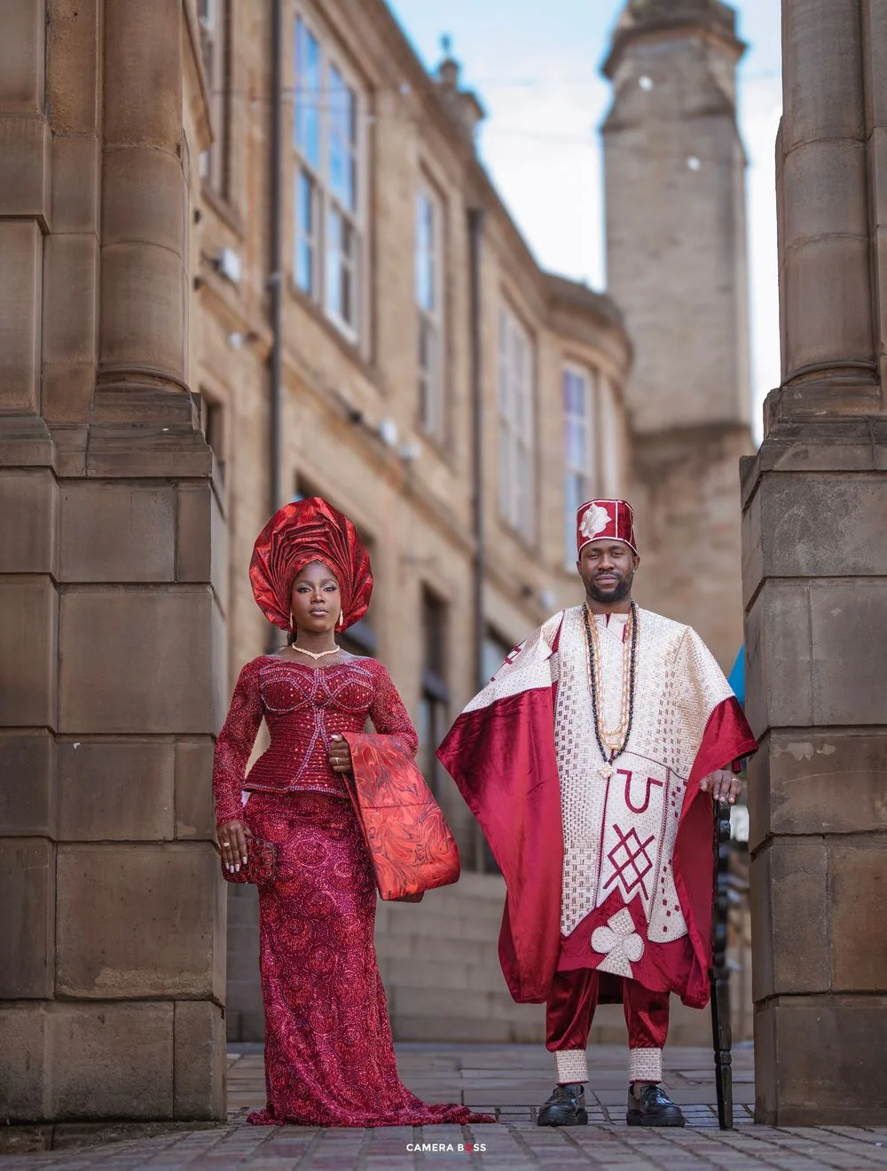 Nigerian couple in traditional Yoruba wedding attire posing between stone columns on a historic cobblestone street.