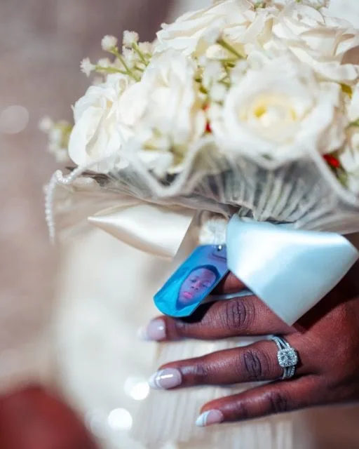 A close-up of hands holding a white wedding bouquet with white roses, pearls, and a decorative blue tag.