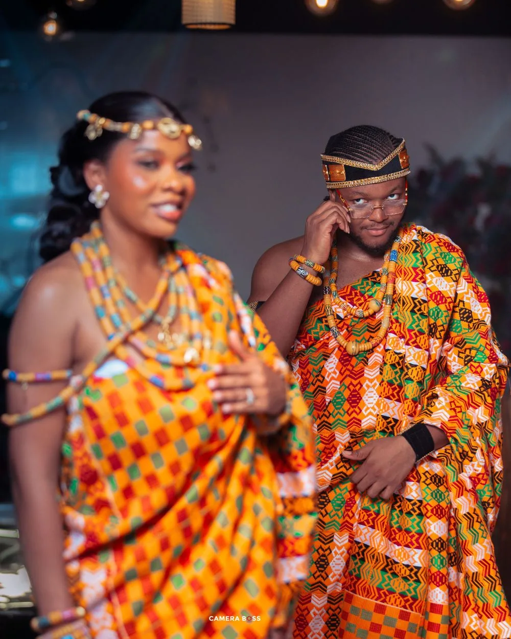 Traditional African wedding celebration with two people wearing vibrant orange kente cloth and colorful cultural attire.