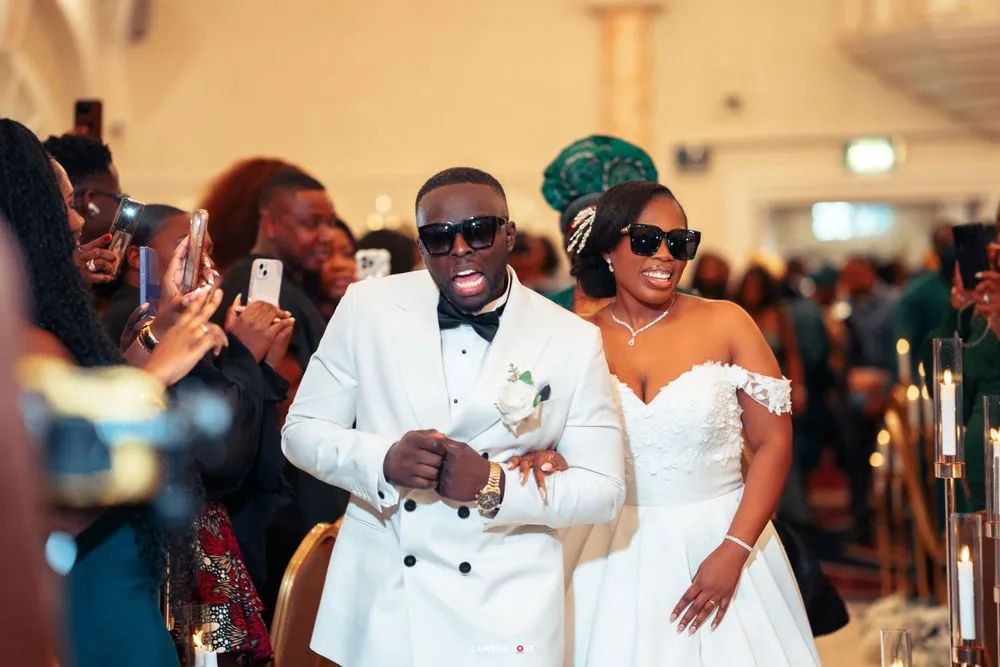 Wedding celebration showing a couple in white formal attire walking together at their reception while guests take photos.