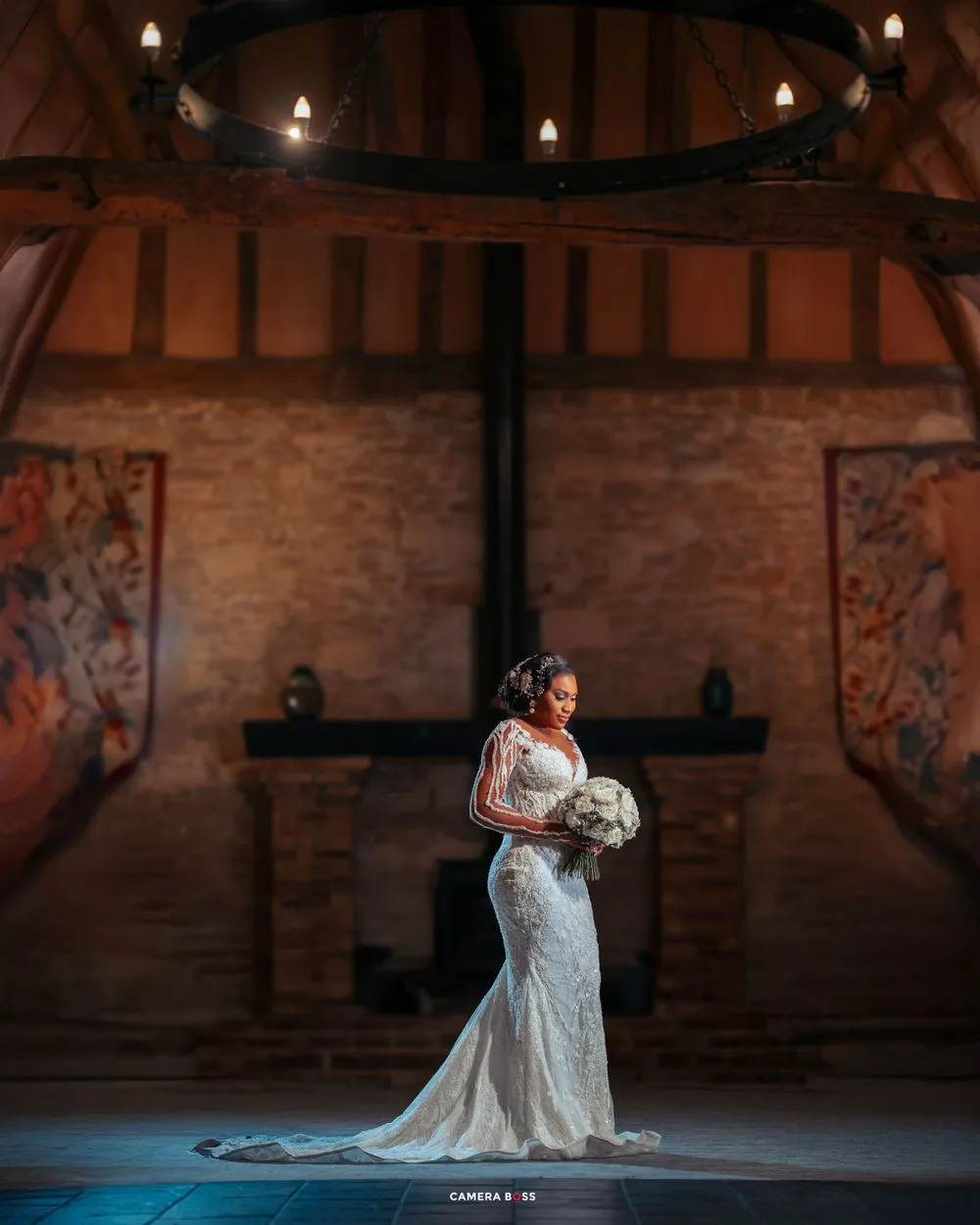 A bride in a fitted lace wedding gown poses elegantly in a rustic barn venue with exposed brick walls and chandelier lighting.