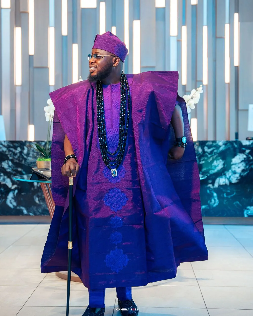 Traditional Nigerian fashion ensemble featuring a dramatic purple agbada robe with blue accents and beaded necklaces.