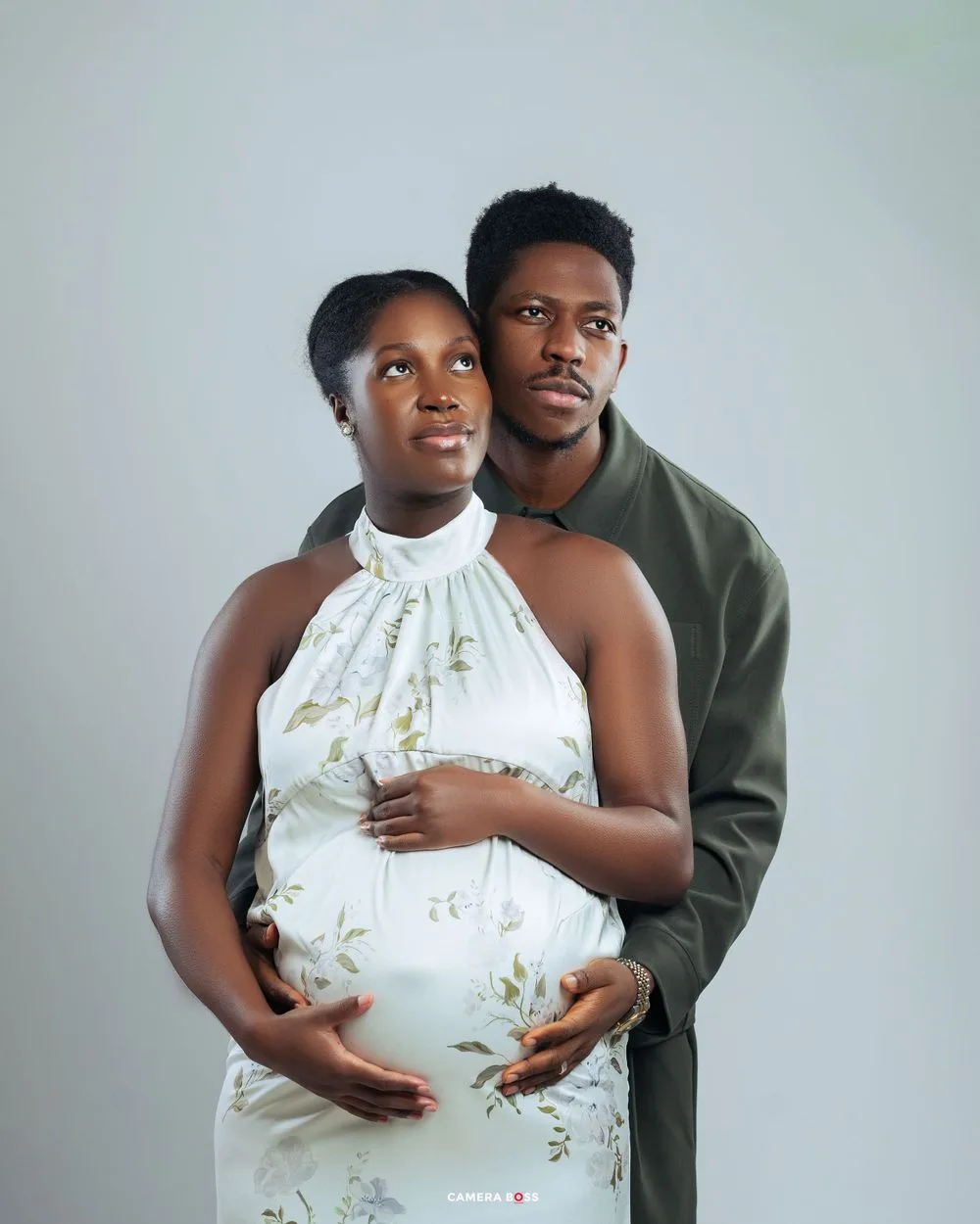 A couple in a maternity photoshoot with the pregnant woman wearing a white floral dress and the partner in a green sweater.