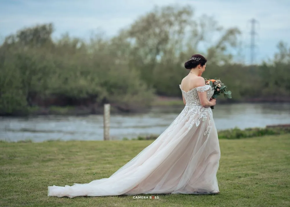 A flowing white wedding dress with lace details and sheer overlay billows in the breeze by a peaceful riverside setting.