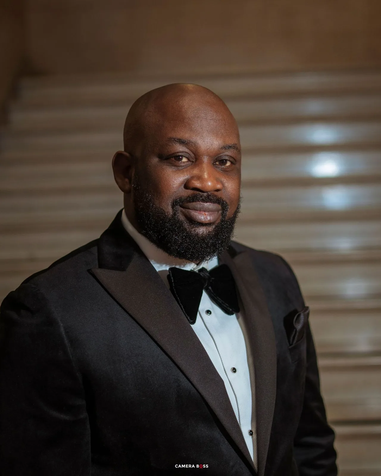 Elegant man in black tuxedo with bow tie smiling confidently on marble staircase background.