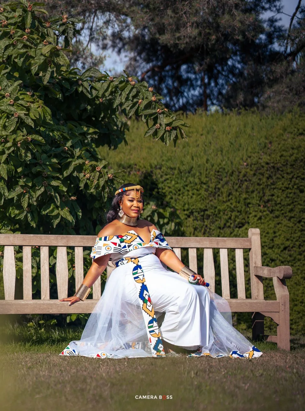 Elegant white dress with African print sash adorns someone seated gracefully on a wooden garden bench in sunlit setting.