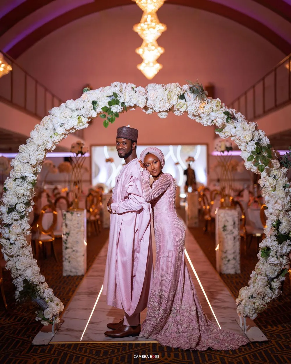 Romantic wedding moment captured under a floral arch with soft pink lighting and white rose decorations.